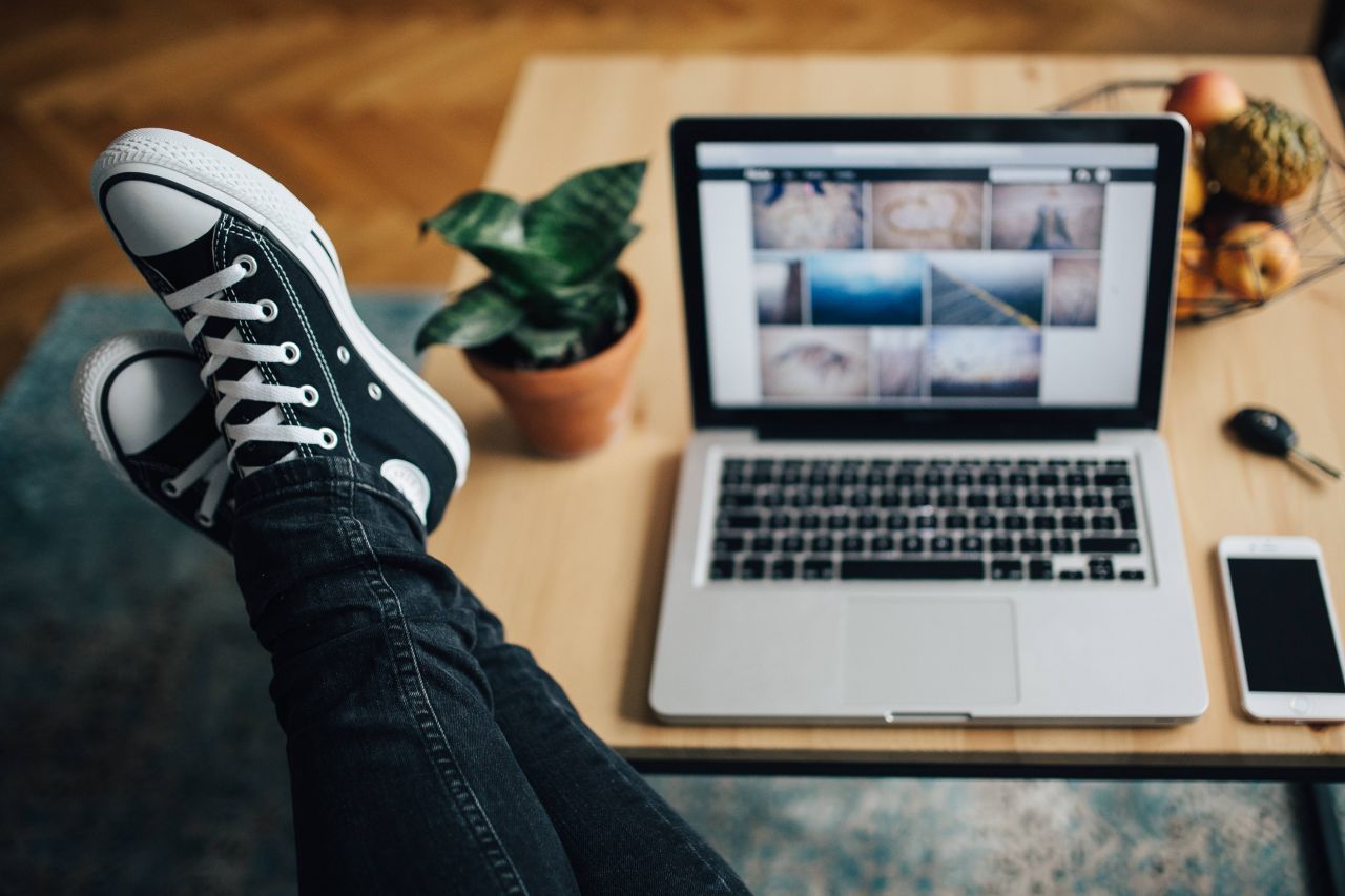 Eine Lehrperson mit den Füssen auf dem Schreibtisch neben einem silbernen Laptop — entspannt, in Sneakers und Jeans. Berichte in Stunden, nicht an Wochenenden.