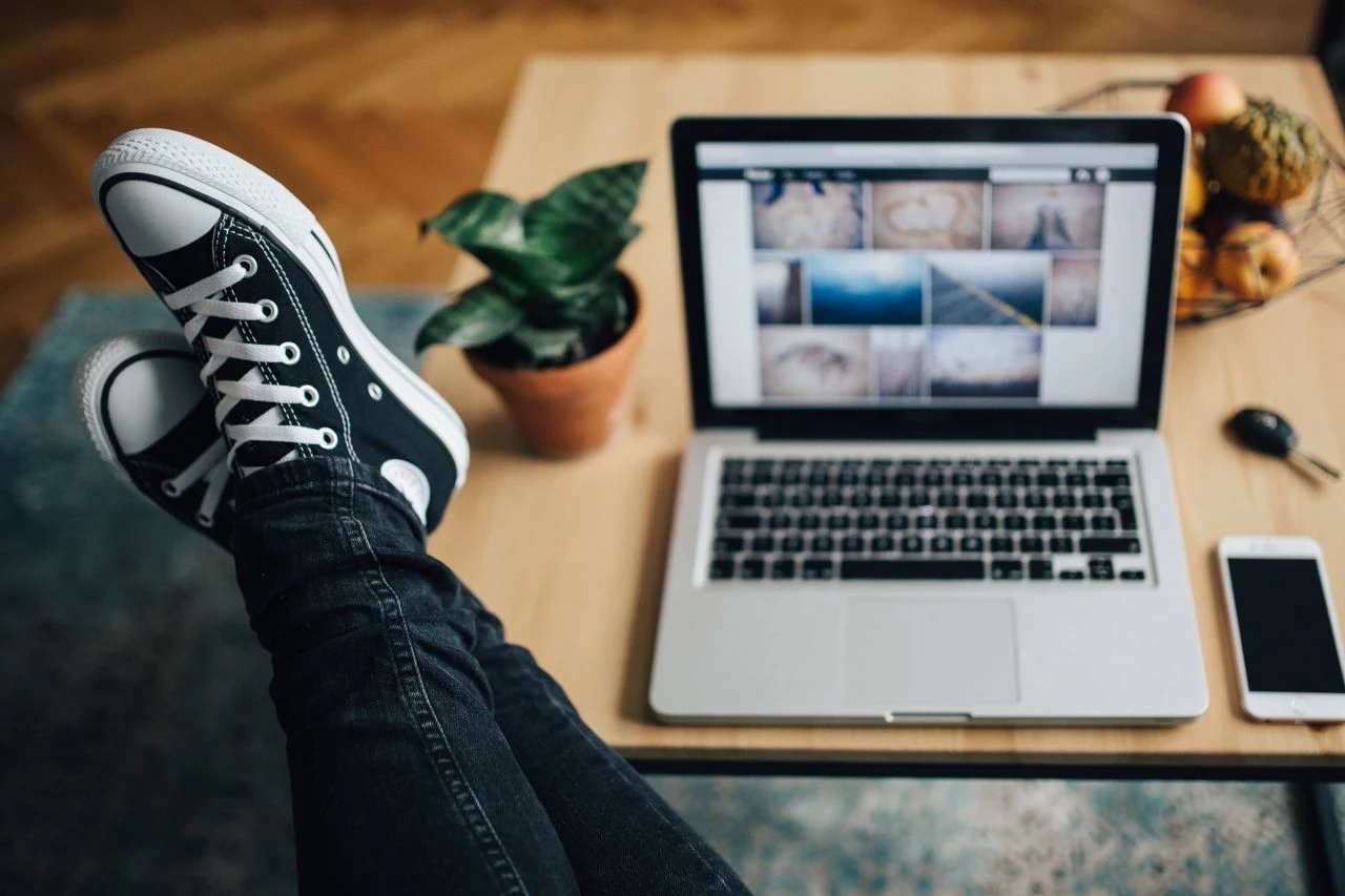 Eine Lehrperson mit den Füssen auf dem Schreibtisch neben einem silbernen Laptop – entspannt, in Sneakers und Jeans.